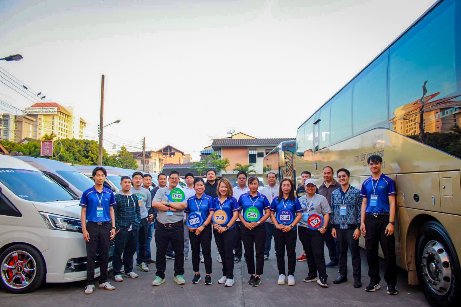 Asian Plus Travel team holding numbered van signs between touring minibuses and a coach
